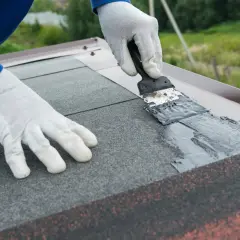 Ręce w białych rękawiczkach uszczelniają dach ciemną masą za pomocą łopatki.
