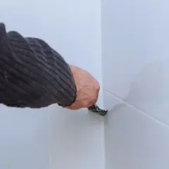 Ręka w swetrze usuwa narzędziem smugi po silikonie z białych płytek.