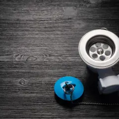 Metalowy syfon pod brodzik z niebieskim korkiem i łańcuszkiem na ciemnym, drewnianym tle.